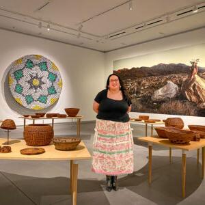梅兰达·罗伯茨站在一圈卡韦拉篮子的中间，篮子放在弯曲的桌子上. 背景是一个由易拉罐制成的篮子和一张沙漠风景的照片.